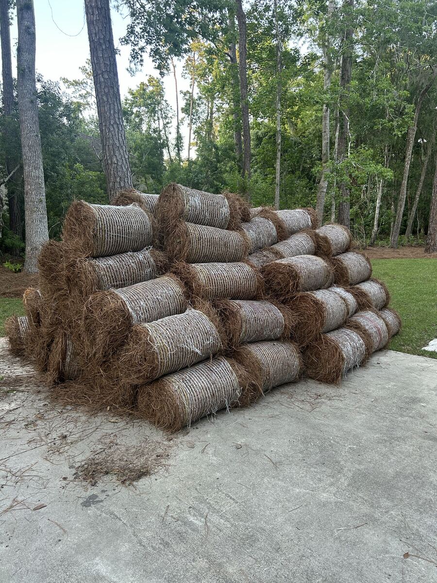 Round bales of pine straw wrapped in netting, stacked several high on a concrete pad with woods behind