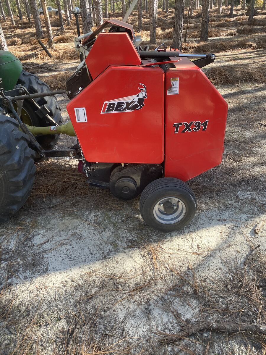 A round baler hitched behind a tractor on the floor of a pine stand