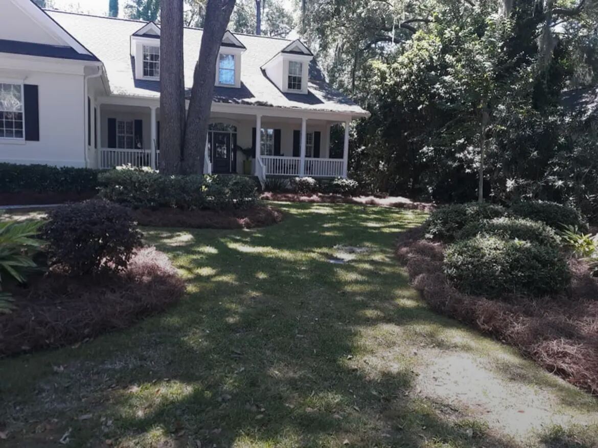 A white house with a covered porch, with pine straw beds curving along both sides of the front lawn